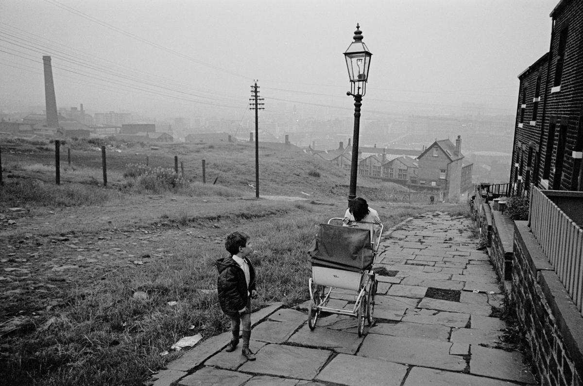 A woman pushes a pram up an uneven pavement with her son, with the Bradford cityscape in the background