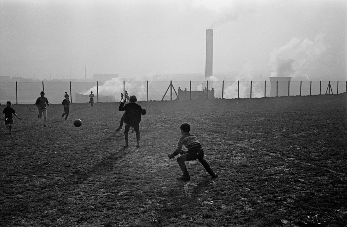 A group of boys play football on a patch of grassland overlooking a power station