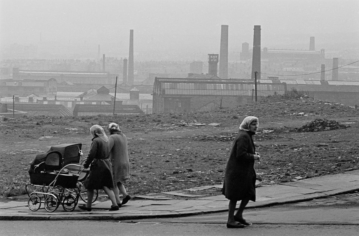 Mothers walk their prams by a patch of waste land overlooking Bradford's industrial mills